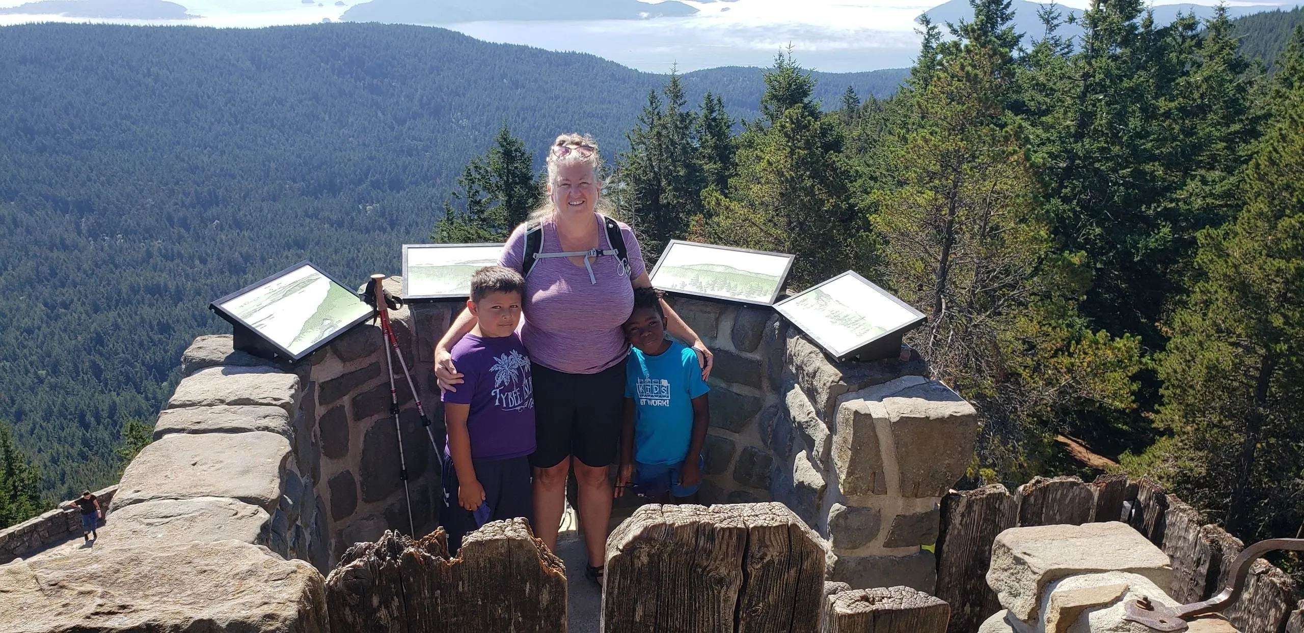 Jill smiling at a stone lookout high above forested islands, with two children beside her.